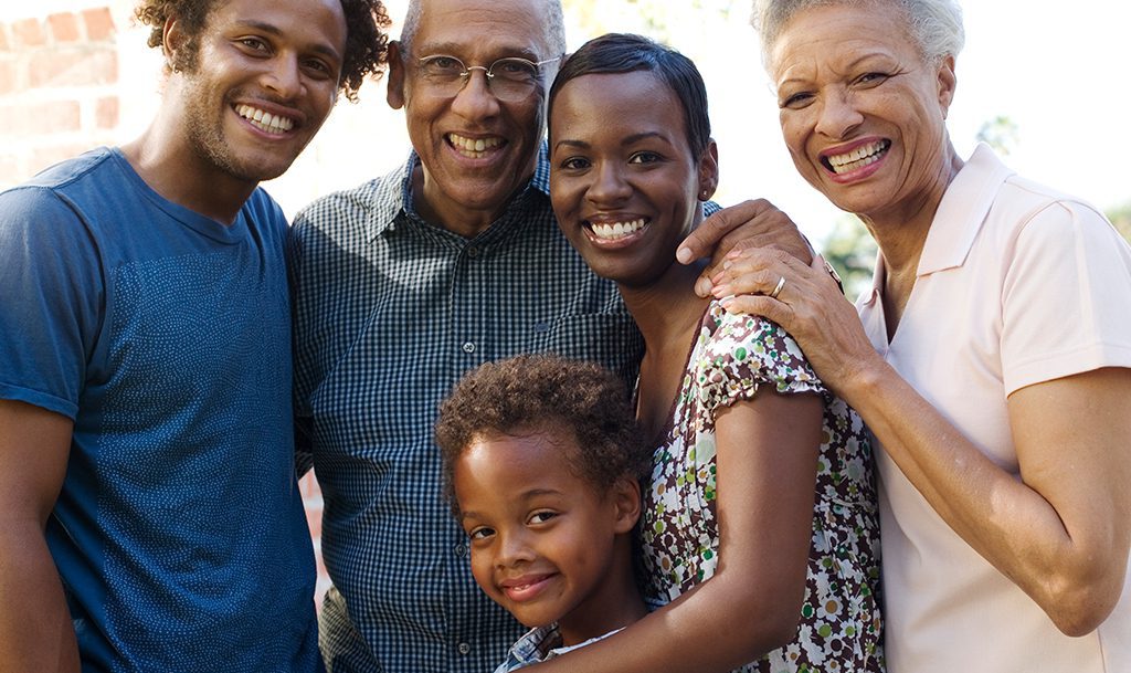 An African American family of four smiles joyfully together, posing for a cheerful photo in a bright outdoor setting.